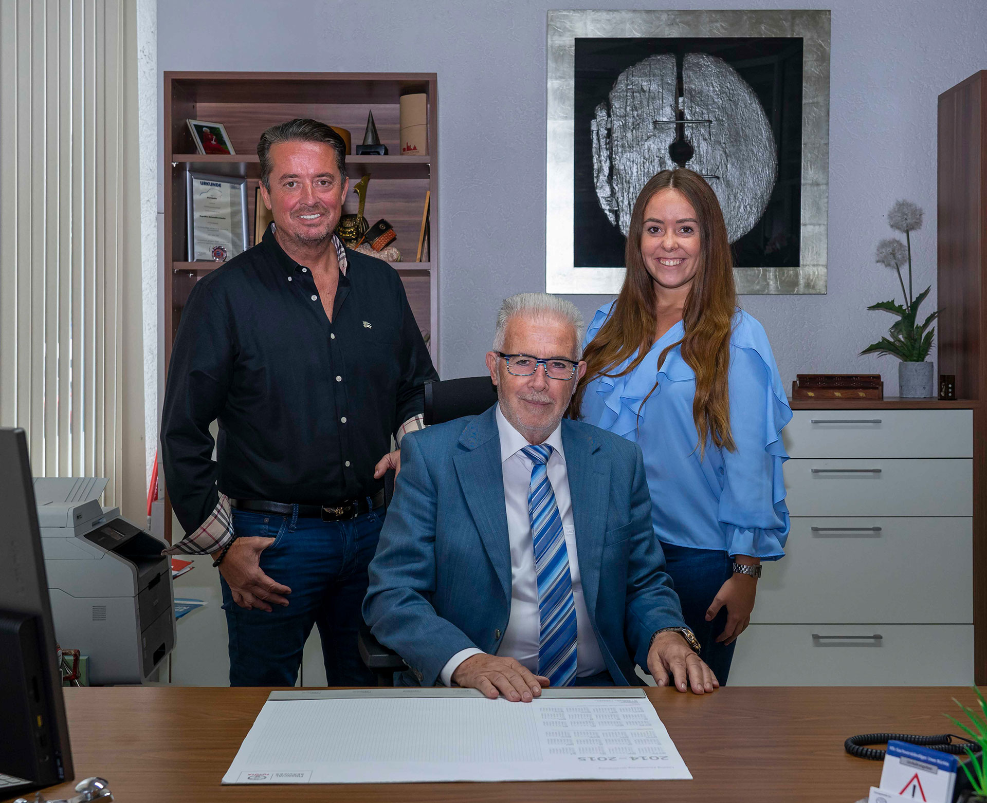 Uwe und Vanessa Bürkle stehen hinter Heinz Bürkle an seinem Schreibtisch im Autohaus.